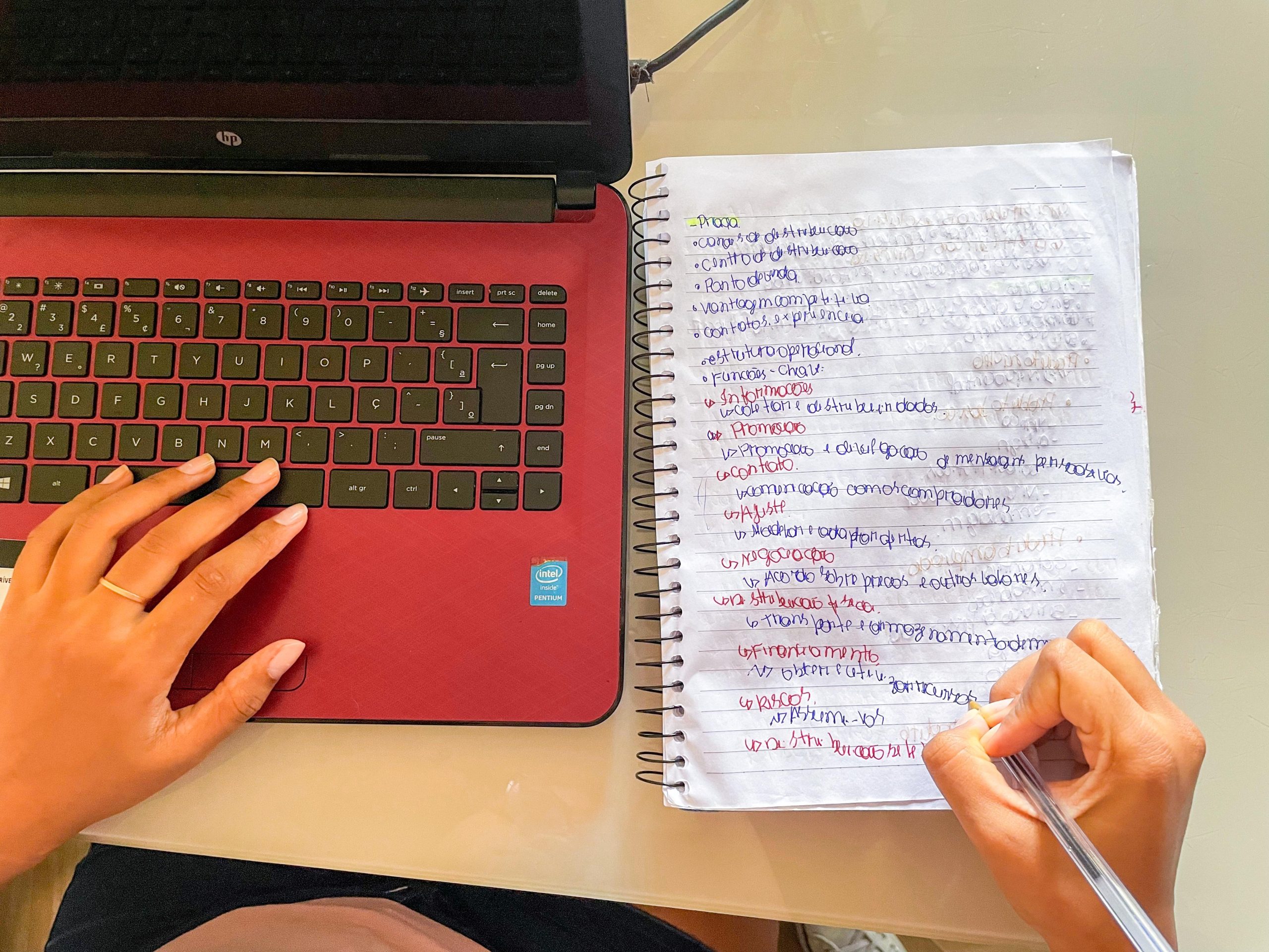 Laptop vermelho e preto. Com uma mão esquerda em cima e a mão direita escreve em um caderno.